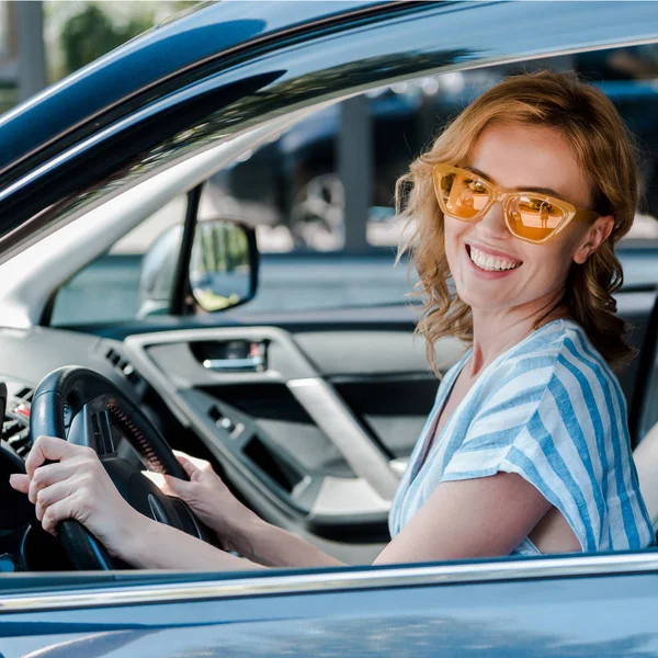 Femme heureuse en lunettes de soleil jaunes tenant volant dans la voiture — Photo de stock