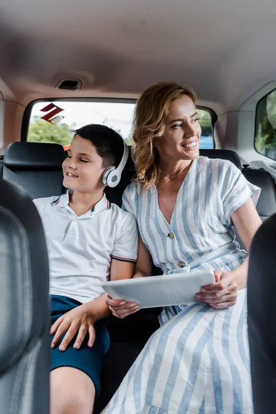 Selektiver Fokus des fröhlichen Kindes im Kopfhörer nahe der Mutter mit digitalem Tablet im Auto — Stockfoto