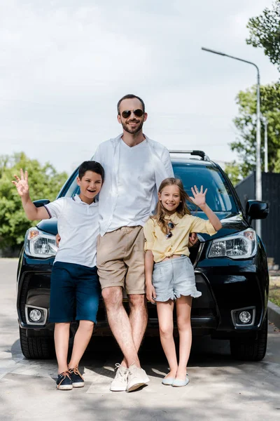 Padre feliz en gafas de sol de pie con los niños agitando las manos cerca del coche - foto de stock