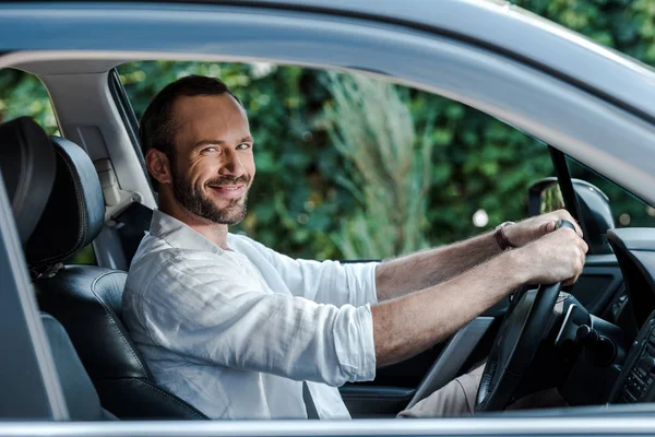 Enfoque selectivo de hombre barbudo feliz sonriendo y mirando a la cámara mientras conduce el coche - foto de stock