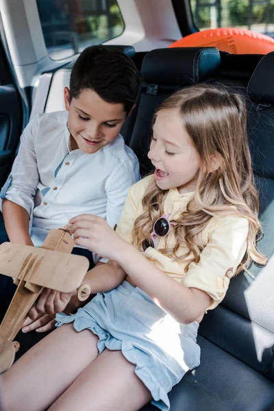 Happy sister and brother playing with wooden toy biplane in car — Stock Photo
