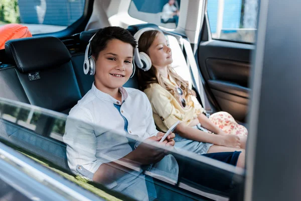 Селективный фокус мальчика с помощью смартфона и прослушивания музыки в наушниках рядом с сестрой в машине — стоковое фото