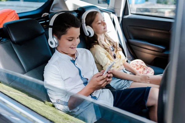 Selective focus of boy using smartphone and listening music in headphones near sister with closed eyes in car — Stock Photo