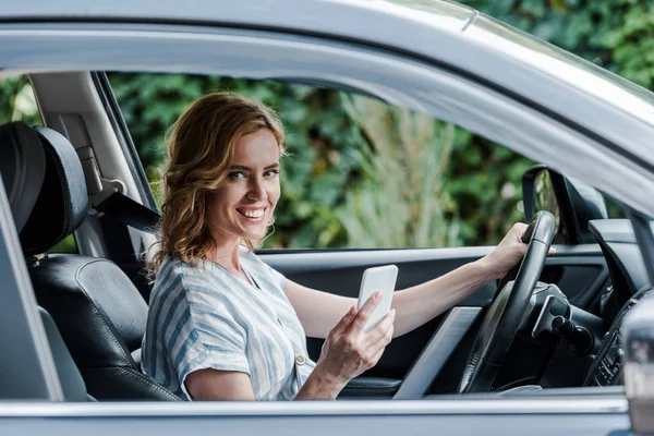 Enfoque selectivo de la mujer sonriente utilizando el teléfono inteligente mientras conduce el coche - foto de stock