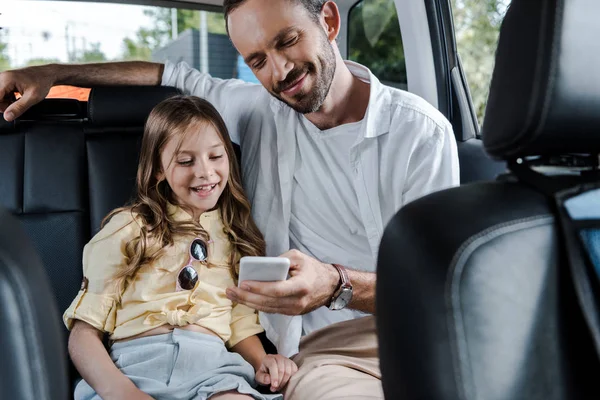 Selective focus of happy man using smartphone near daughter in car — Stock Photo