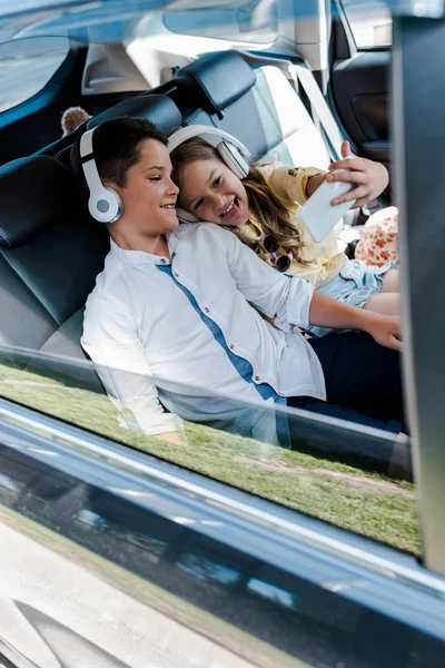 Selective focus of cheerful kid in headphones taking selfie with brother in car — Stock Photo