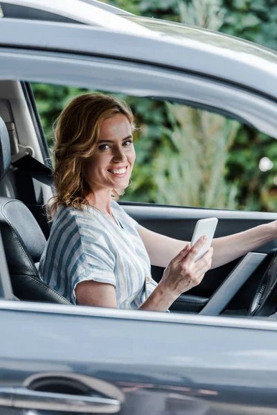 Selektiver Fokus der glücklichen Frau, die ihr Smartphone im Auto hält — Stockfoto