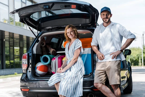 Schöner Mann mit Mütze, der mit der Hand auf der Hüfte neben attraktiver Frau und Auto steht — Stockfoto