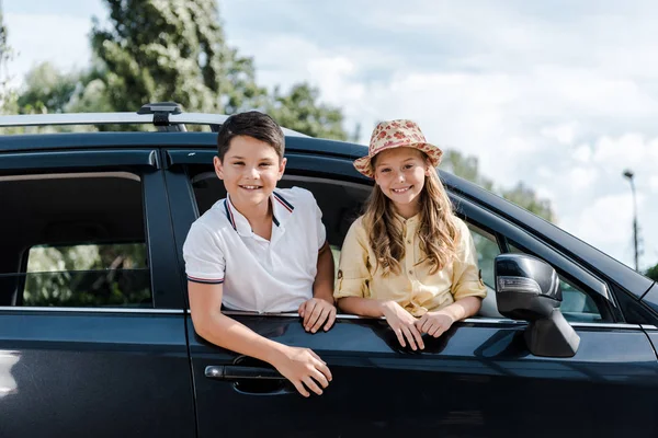 Sorella felice in cappello di paglia e fratello guardando la fotocamera dai finestrini dell'auto — Foto stock