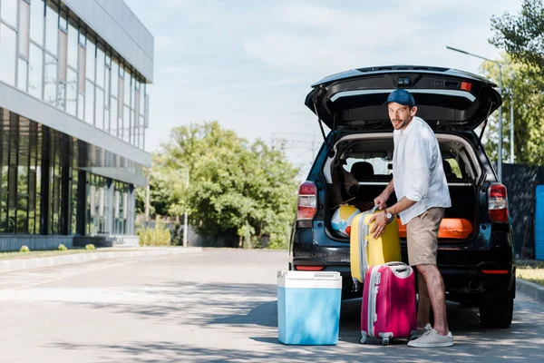 Homme barbu gai en casquette debout près des bagages et de la voiture — Photo de stock
