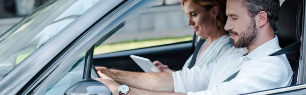 Панорамный снимок мужчины за рулем автомобиля рядом с женщиной с помощью цифрового планшета — стоковое фото