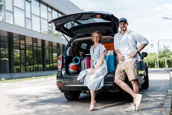 Homme gai en chapeau debout avec la main sur la hanche près de la femme et de la voiture — Photo de stock