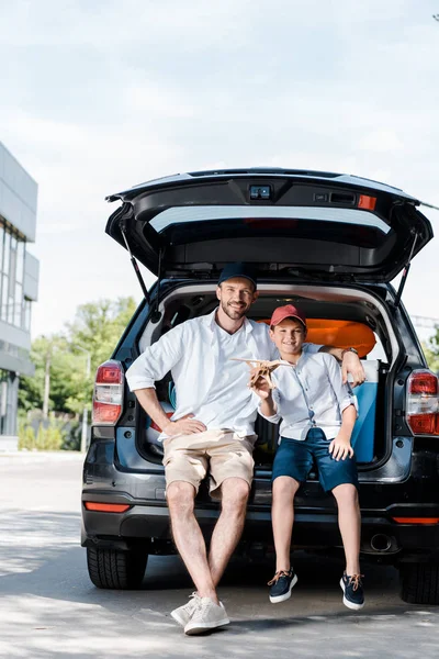 Fröhlicher Vater sitzt neben Sohn mit hölzernem Doppeldecker neben Auto — Stockfoto