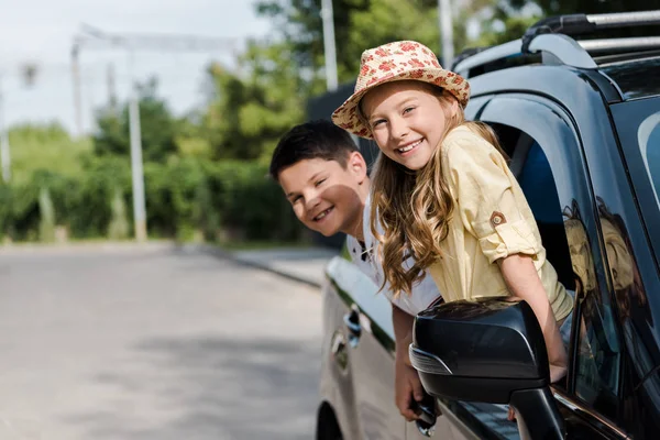 Selective focus of happy sister and brother looking at camera from car windows — Stock Photo