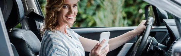 Tiro panorâmico de mulher feliz segurando smartphone no carro — Fotografia de Stock