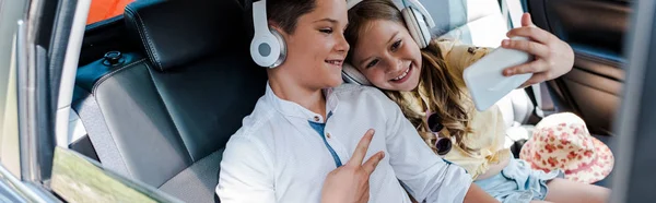 Panoramic shot of kid showing peace sign near sister taking selfie in car — Stock Photo