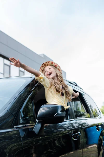 Blick auf ein glückliches Kind mit Strohhut und ausgestreckten Händen im Autofenster — Stockfoto