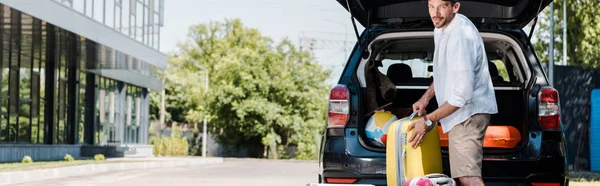 Panoramaaufnahme eines gut gelaunten bärtigen Mannes mit Mütze, der in der Nähe von Gepäck und Auto steht — Stockfoto