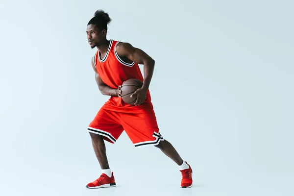 Jugador de baloncesto afroamericano en ropa deportiva roja sosteniendo pelota sobre fondo gris - foto de stock