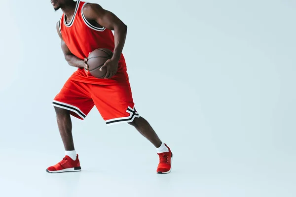 Teilansicht eines afrikanisch-amerikanischen Sportlers in roter Sportbekleidung beim Basketballspielen auf grauem Hintergrund — Stockfoto