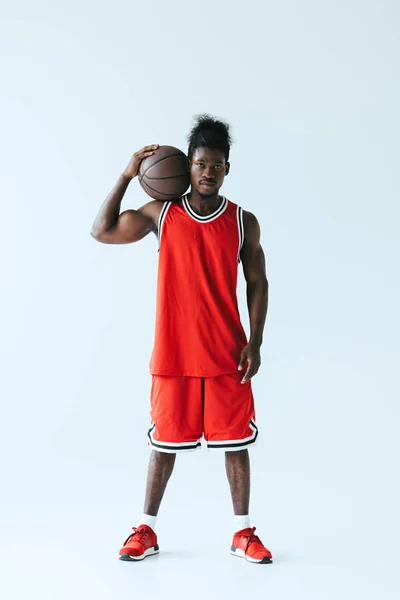 Confiado jugador de baloncesto afroamericano en ropa deportiva roja sosteniendo la pelota y mirando a la cámara sobre fondo gris - foto de stock