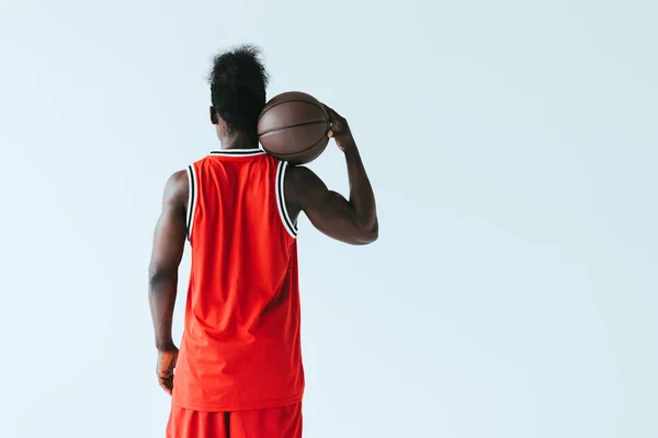 Vista trasera del jugador de baloncesto afroamericano en ropa deportiva roja sosteniendo la pelota aislada en gris - foto de stock