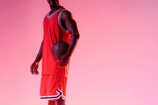 Vista recortada de jugador de baloncesto afroamericano sosteniendo pelota sobre fondo rosa con gradiente - foto de stock
