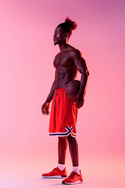Schöner afrikanisch-amerikanischer Basketballspieler, der auf rosa und lila Gradienten-Hintergrund mit Beleuchtung wegschaut — Stockfoto