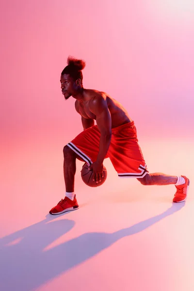 Без сорочки афроамериканський спортсмен з м'язистим торсом грає в баскетбол на рожевому фоні з градієнтом і освітленням — стокове фото
