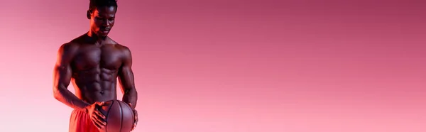 Tiro panorâmico de shirtless, muscular jogador de basquete americano africano segurando bola no fundo rosa escuro com gradiente — Fotografia de Stock