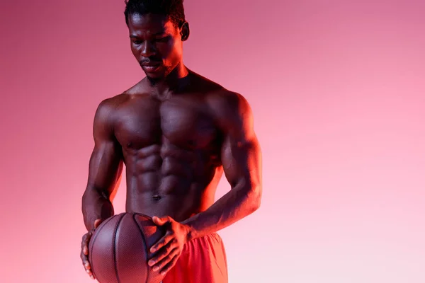 Серьезный африканский американский спортсмен с мускулистым туловищем держит мяч на розовом фоне с градиентом — стоковое фото