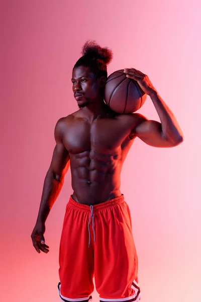 Впевнений, без сорочки афроамериканський спортсмен, дивлячись далеко, тримаючи м'яч на рожевому фоні з градієнтом — стокове фото