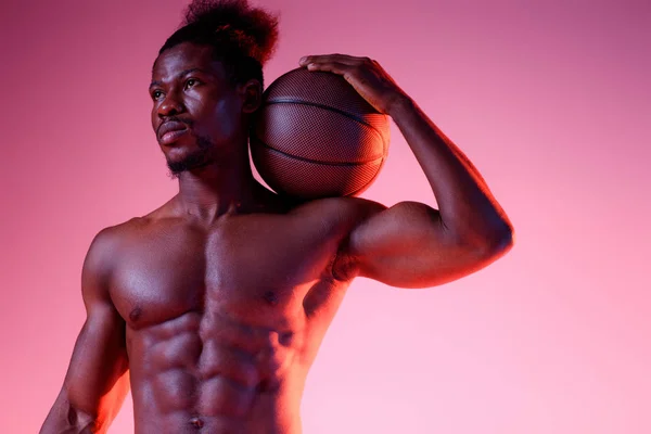 Серьезный африканский американский спортсмен с мускулистым туловищем, держащий мяч и смотрящий в сторону на розовый и фиолетовый градиентный фон — стоковое фото