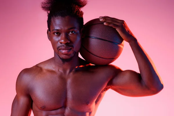 Уверенно, без рубашки африканский баскетболист держит мяч и смотрит на камеру на розовом фоне с градиентом — стоковое фото