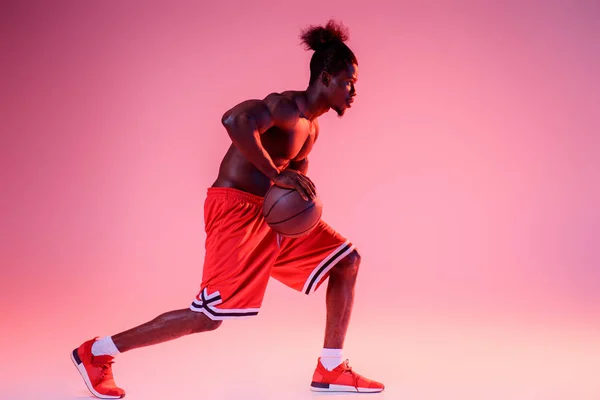 African american sportsman in red short playing basketball on pink and purple gradient background — Stock Photo