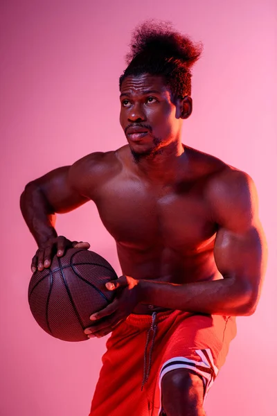 Jugador de baloncesto afroamericano serio sosteniendo la pelota y mirando hacia otro lado en el fondo de degradado rosa y púrpura - foto de stock