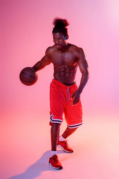 Sportif afro-américain torse nu en short rouge jouant au basket sur fond rose avec dégradé et éclairage — Photo de stock