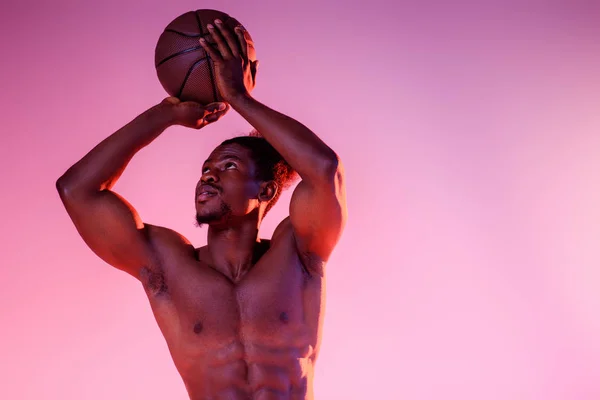 Guapo afroamericano deportista con torso muscular jugando baloncesto en rosa y púrpura gradiente fondo - foto de stock