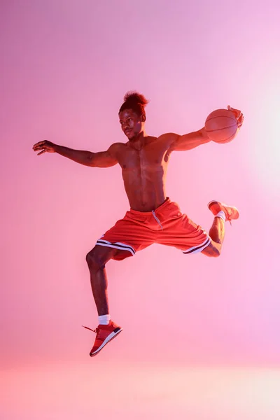 Shirtless african american sportsman in red shorts jumping while playing basketball on pink and purple gradient background — Stock Photo