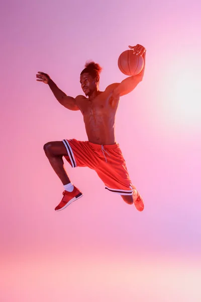 Handsome, muscular african american sportsman in red shorts jumping while playing basketball on pink and purple gradient background — Stock Photo