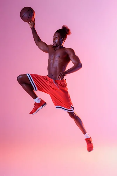 Athletic african american sportsman in red shorts playing basketball on pink and purple gradient background — Stock Photo
