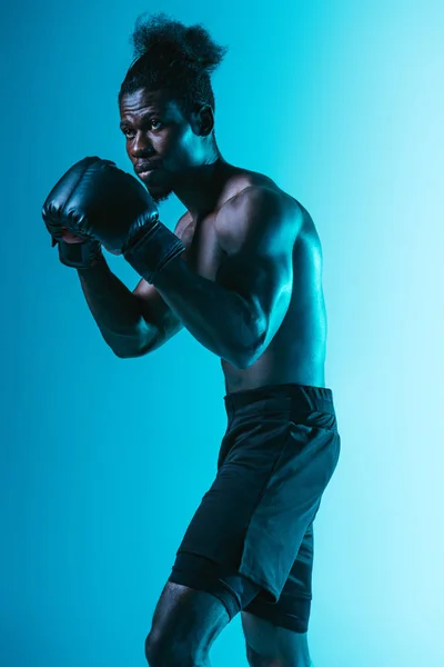 Selbstbewusster afrikanisch-amerikanischer Sportler mit muskulösem Oberkörper beim Boxen auf blauem Hintergrund — Stockfoto