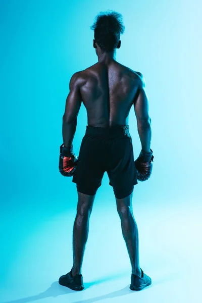 Back view of shirtless, muscular african american sportsman in boxing gloves on blue background — Stock Photo