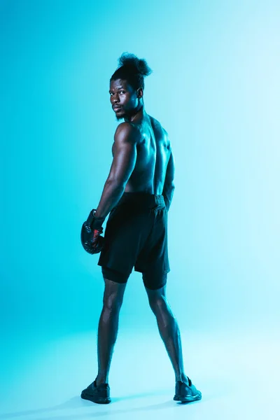 Серьезный африканский американский спортсмен в боксёрских перчатках смотрит в камеру на синем фоне — стоковое фото