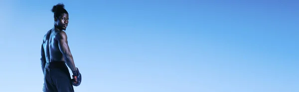 Панорамный снимок безрубашечного, мускулистого африканского боксера, смотрящего на камеру на синем фоне — стоковое фото