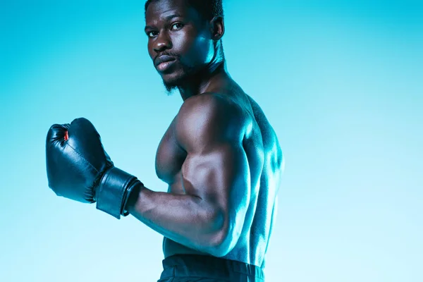 Ernster afrikanisch-amerikanischer Boxer mit muskulösem Oberkörper, der in die Kamera auf blauem Hintergrund blickt — Stockfoto