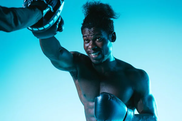 Musculoso boxeador afroamericano haciendo ejercicio con entrenador sobre fondo azul - foto de stock