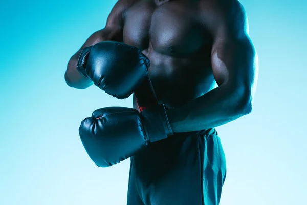 Corte vista de shirtless, muscular afro-americano desportista em luvas de boxe no fundo azul — Fotografia de Stock