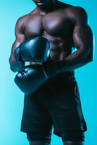 Partial view of shirtless, muscular african american sportsman in boxing gloves on blue background — Stock Photo