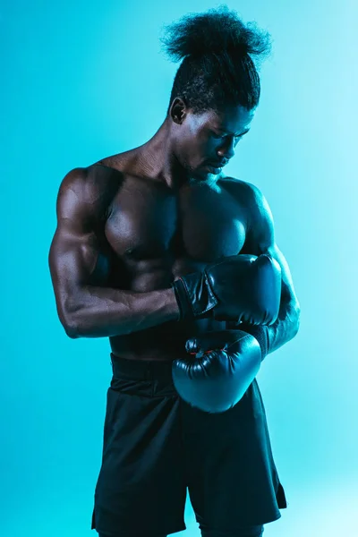 Sério, muscular afro-americano desportista em luvas de boxe posando no fundo azul — Fotografia de Stock
